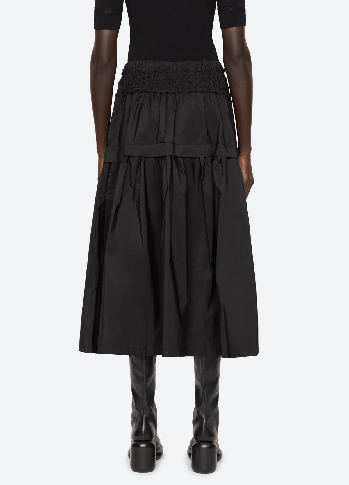 A person wearing the Belen Taffeta Smocked Tiered Skirt by Sea New York, paired with knee-high black boots. The outfit is completed with a black short-sleeve top, and the person stands against a plain, light-colored background.