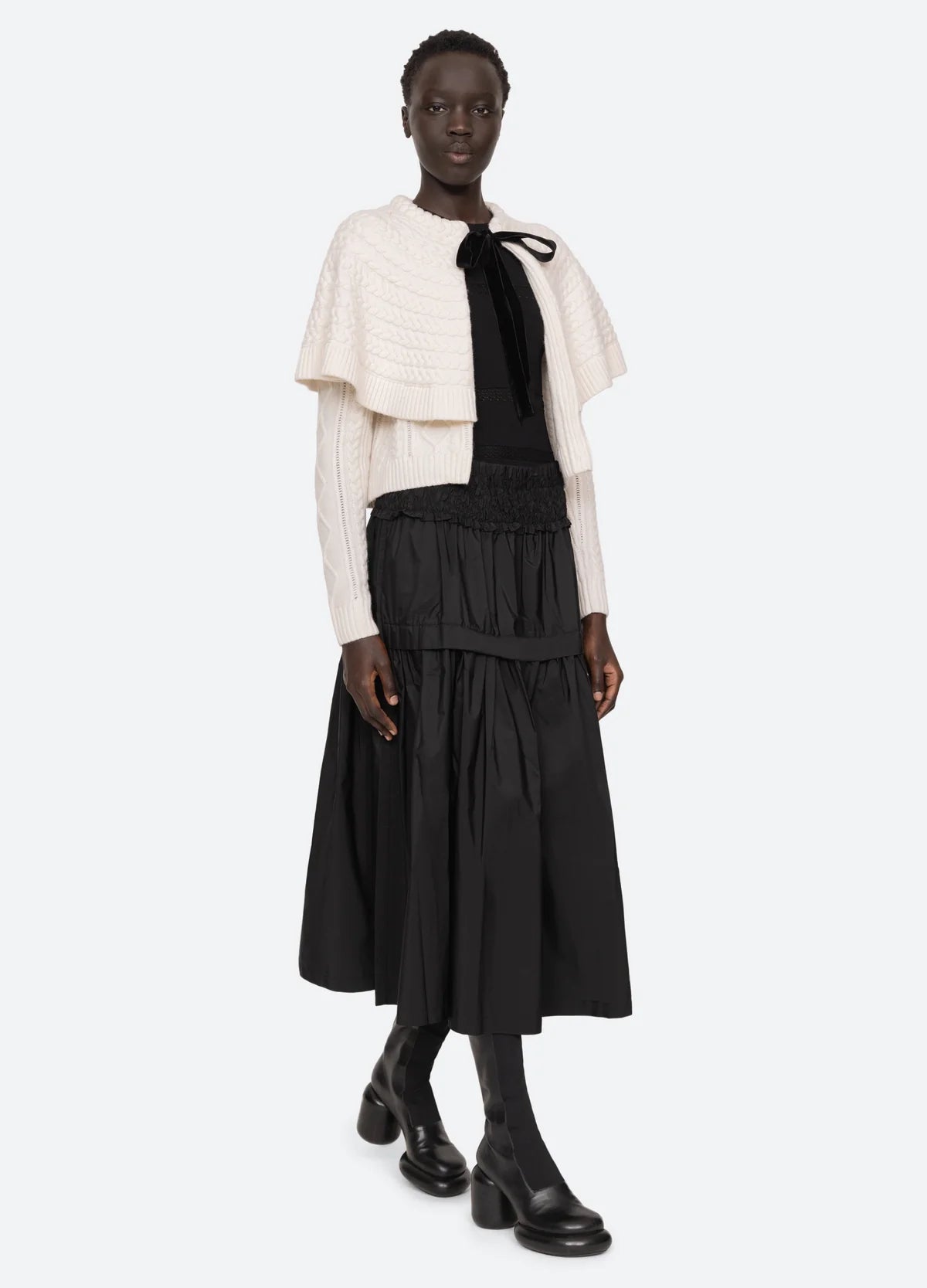 A person is showcasing a stylish ensemble with a cream knit cape and the Belen Taffeta Smocked Tiered Skirt from Sea New York, featuring a flowing black design. Paired with black boots, they stand against a plain white background, expertly combining textures for a chic layered look.