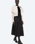 A person is showcasing a stylish ensemble with a cream knit cape and the Belen Taffeta Smocked Tiered Skirt from Sea New York, featuring a flowing black design. Paired with black boots, they stand against a plain white background, expertly combining textures for a chic layered look.