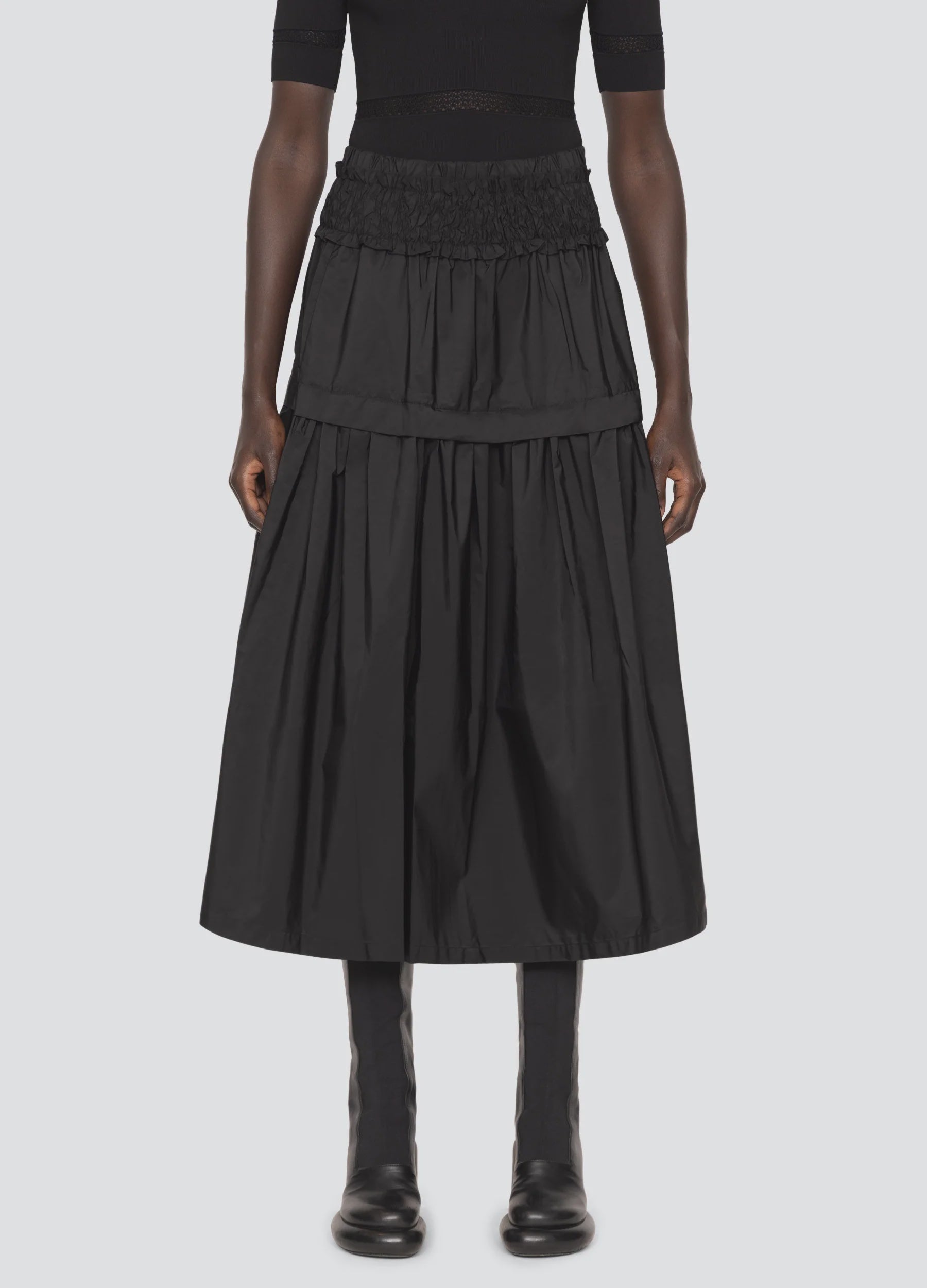 A person stands against a light background, showcasing the Belen Taffeta Smocked Tiered Skirt by Sea New York, featuring a black smocked waistband. The tiered midi silhouette is complemented by a matching black top and boots.