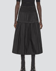 A person stands against a light background, showcasing the Belen Taffeta Smocked Tiered Skirt by Sea New York, featuring a black smocked waistband. The tiered midi silhouette is complemented by a matching black top and boots.