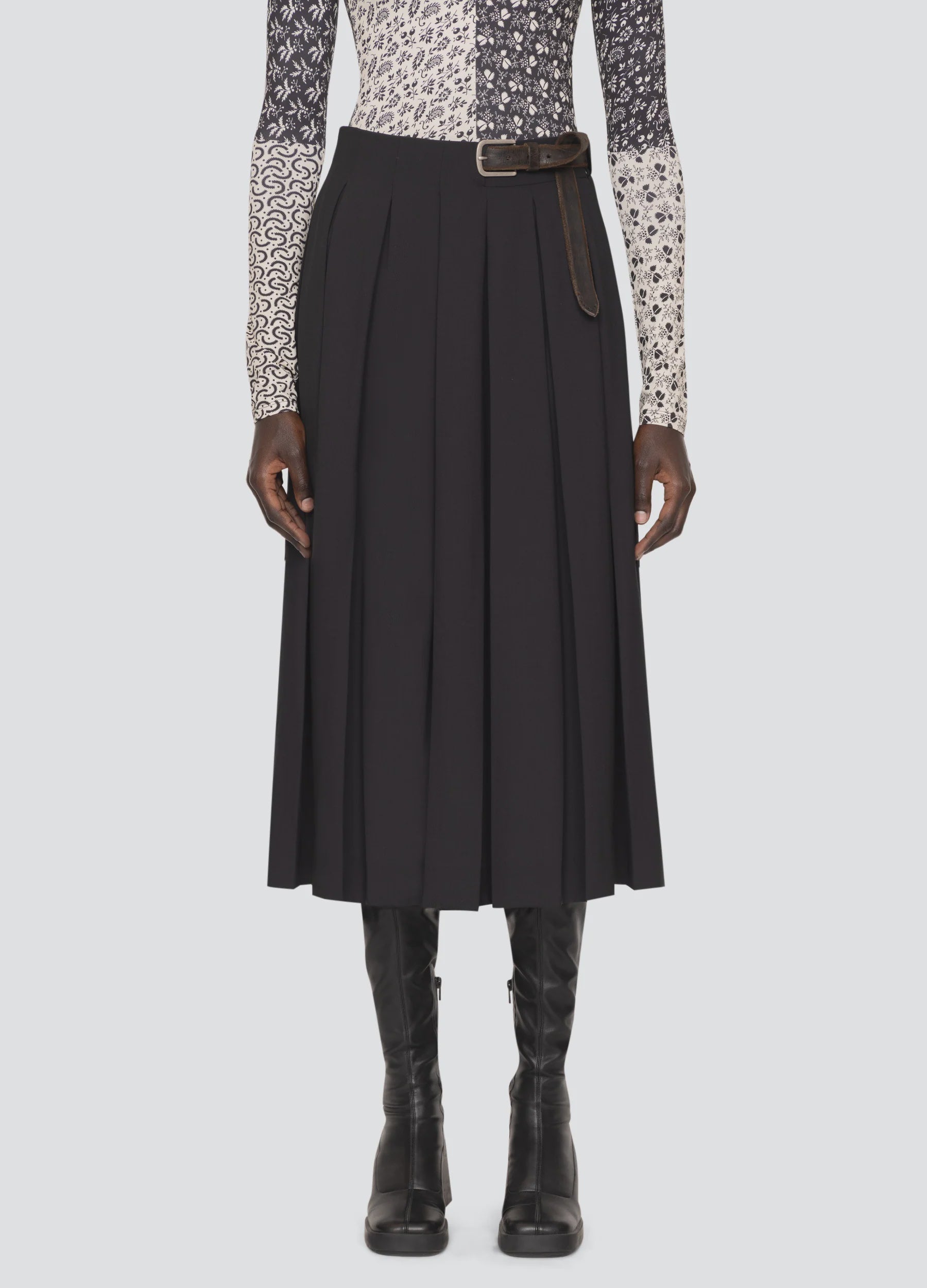 A person is standing in a Sea New York Hallie Suiting Skirt, which is a black pleated midi skirt with a high-rise waist and belt. They are wearing knee-high black boots and a long-sleeve top featuring mixed patterns in shades of black, white, and gray against a plain white background.