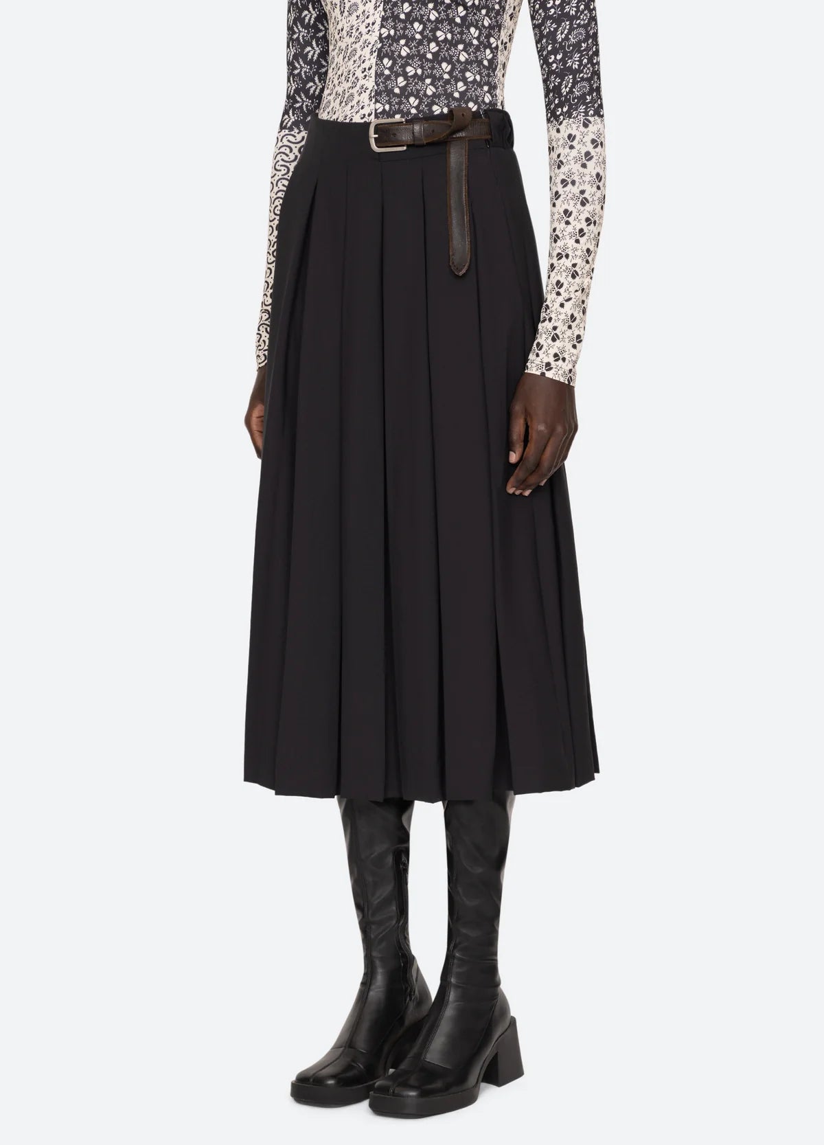 A person wearing a long-sleeved patterned top, the Hallie Suiting Skirt by Sea New York featuring a wool fabric with pleated detailing and a high-rise waist, paired perfectly with knee-high black boots.