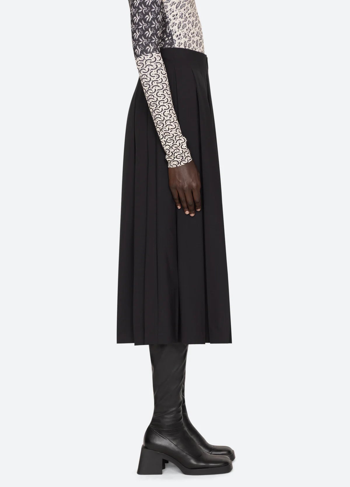 A person is shown in profile wearing a patterned long-sleeve top, the Hallie Suiting Skirt by Sea New York which features a high-waisted fit and pleated detailing, and knee-high black heeled boots. The setting is against a plain, light-colored background.