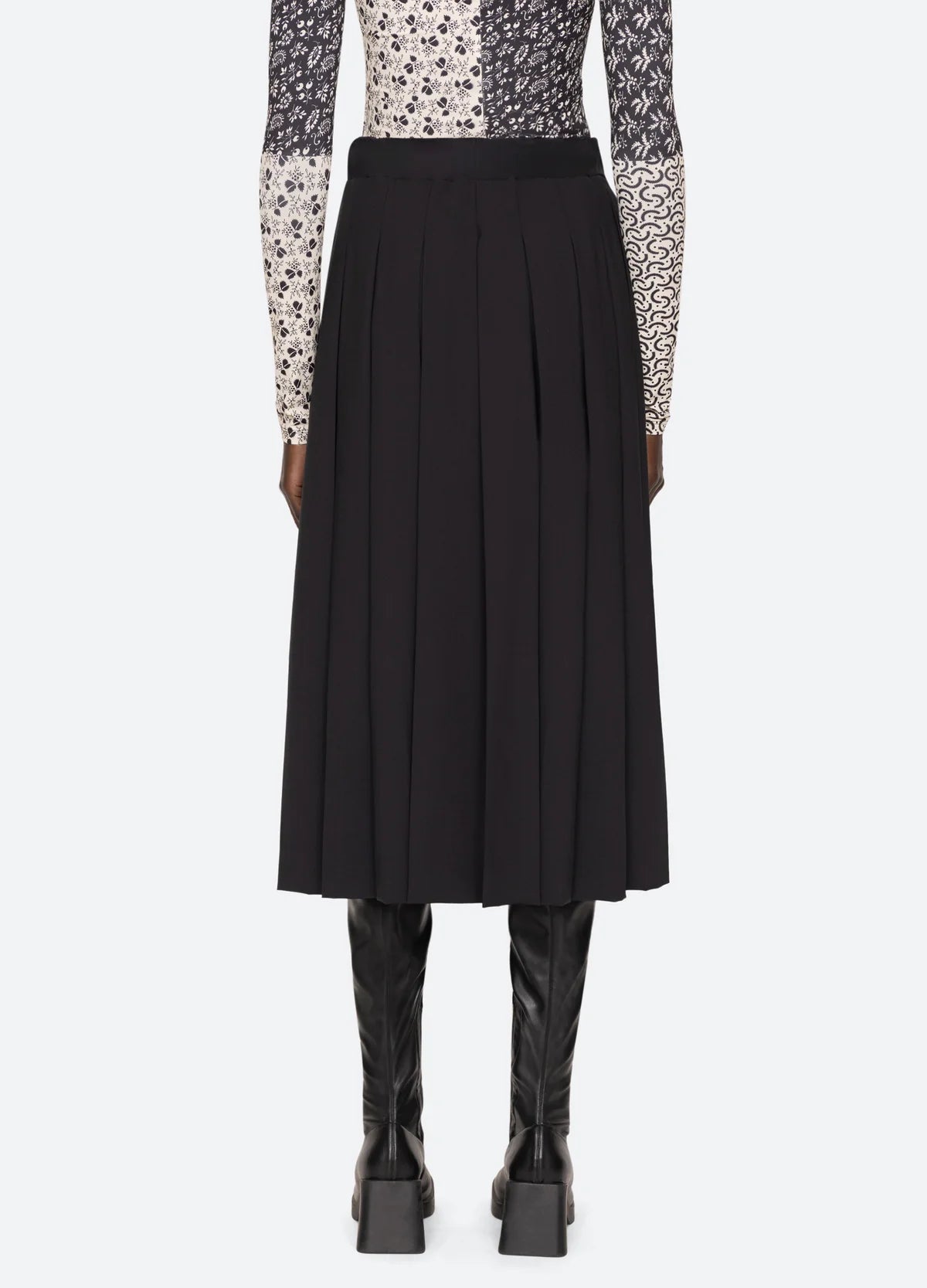 A person wearing a patterned long-sleeve top and the Hallie Suiting Skirt by Sea New York, a black wool midi skirt featuring pleated detailing accentuating its high-rise waist, stands with their back to the camera against a light gray background, paired with knee-high black leather boots.