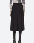 A person wearing a patterned long-sleeve top and the Hallie Suiting Skirt by Sea New York, a black wool midi skirt featuring pleated detailing accentuating its high-rise waist, stands with their back to the camera against a light gray background, paired with knee-high black leather boots.