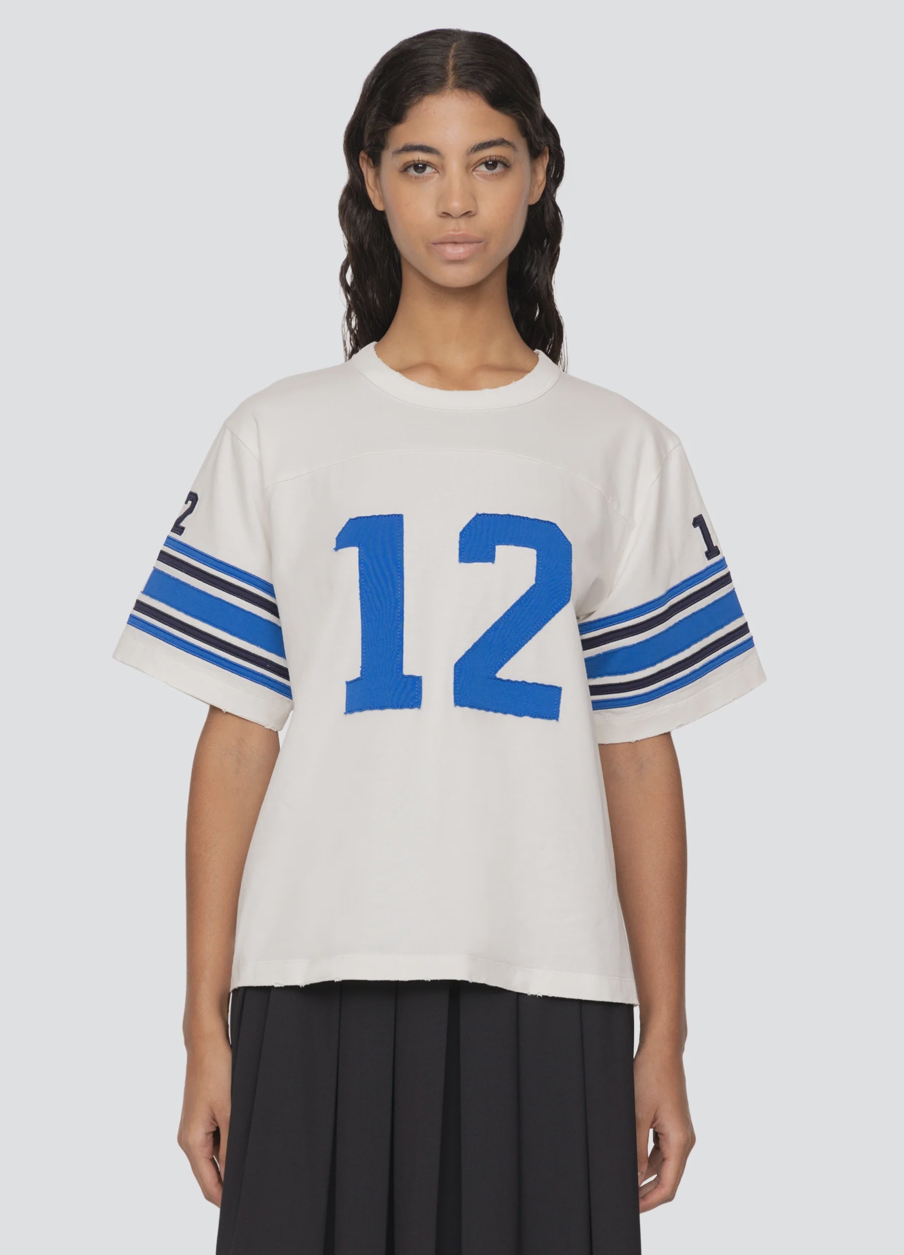 A person stands wearing Sea New York's Finnley Football Graphic T-Shirt, which showcases a relaxed fit and features the number 12 in blue on the front and sleeves. The white short-sleeve shirt is adorned with blue stripes on the sleeves and is crafted from 100% cotton. The background is light gray.