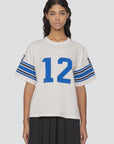 A person stands wearing Sea New York's Finnley Football Graphic T-Shirt, which showcases a relaxed fit and features the number 12 in blue on the front and sleeves. The white short-sleeve shirt is adorned with blue stripes on the sleeves and is crafted from 100% cotton. The background is light gray.