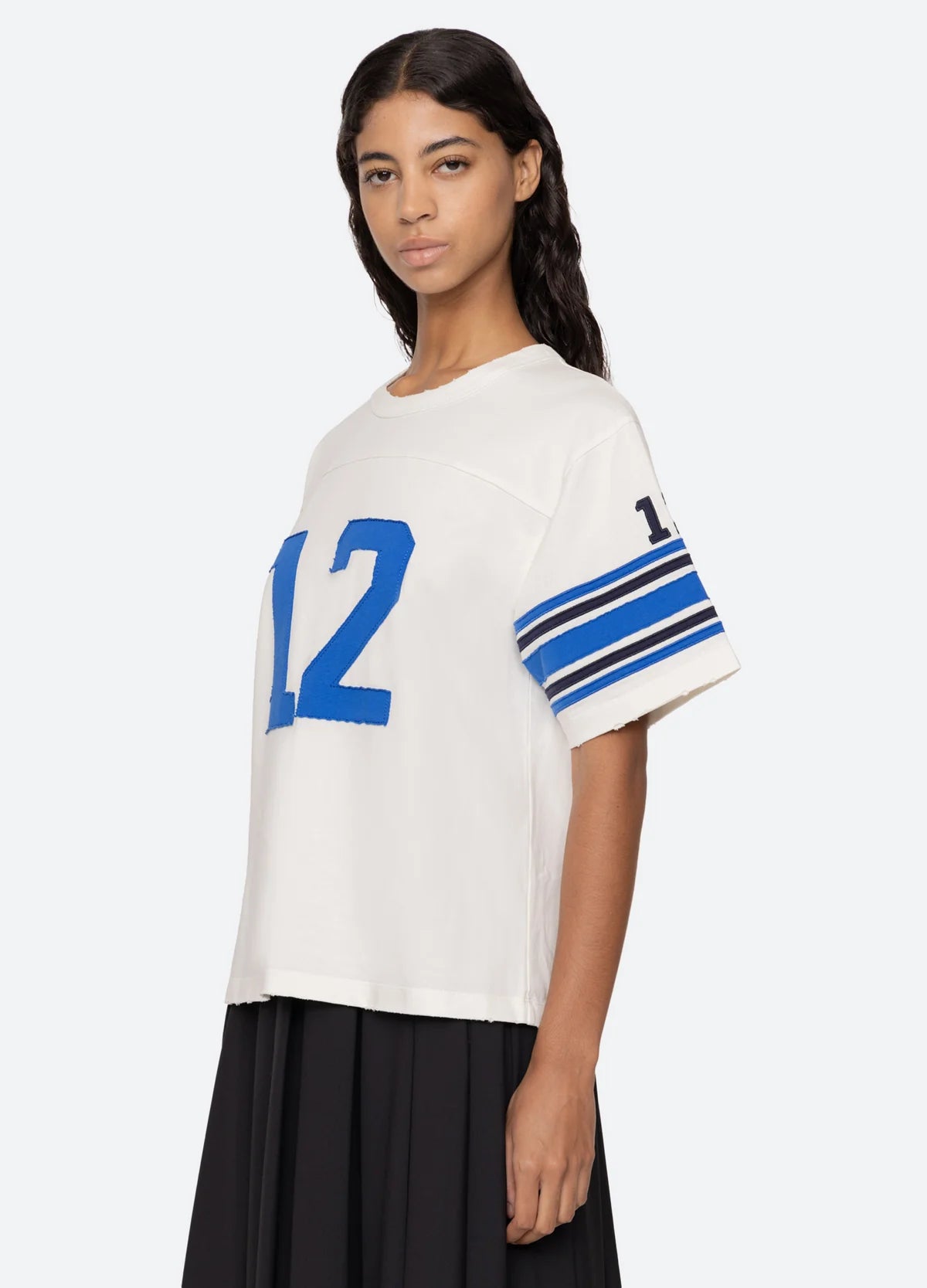 A person wearing the Sea New York Finnley Football Graphic T-Shirt, which is white with the number 12 in blue on the front and sleeves, stands against a light background, showcasing a sporty silhouette. The T-shirt features blue stripes on the sleeves and complements their long wavy hair along with a relaxed-fit black skirt.