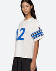 A person wearing the Sea New York Finnley Football Graphic T-Shirt, which is white with the number 12 in blue on the front and sleeves, stands against a light background, showcasing a sporty silhouette. The T-shirt features blue stripes on the sleeves and complements their long wavy hair along with a relaxed-fit black skirt.