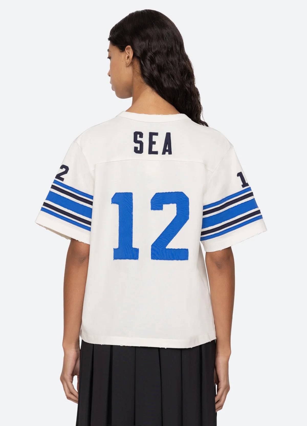 A person with long dark hair is wearing a relaxed fit Finnley Football Graphic T-Shirt by Sea New York, featuring "SEA" and the number "12" in blue on a white background. The sporty design includes sleeves adorned with blue stripes and the number "12" on each arm. They are standing with their back to the camera, reminiscent of a classic Finnley graphic t-shirt style.