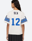 A person with long dark hair is wearing a relaxed fit Finnley Football Graphic T-Shirt by Sea New York, featuring "SEA" and the number "12" in blue on a white background. The sporty design includes sleeves adorned with blue stripes and the number "12" on each arm. They are standing with their back to the camera, reminiscent of a classic Finnley graphic t-shirt style.