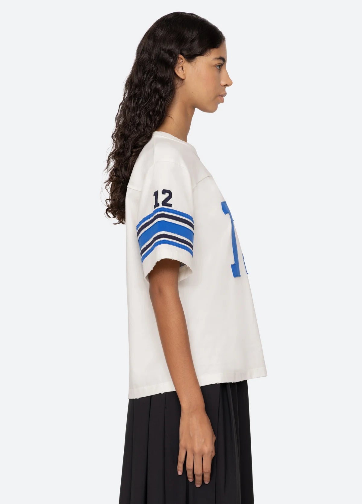 A person with long, curly hair is wearing the Finnley Football Graphic T-Shirt by Sea New York, featuring a relaxed fit and blue stripes, along with the number 12 on the sleeve. They stand in profile against a plain background, complemented by a dark pleated skirt that enhances their laid-back, sporty silhouette.