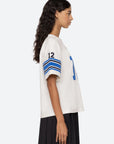 A person with long, curly hair is wearing the Finnley Football Graphic T-Shirt by Sea New York, featuring a relaxed fit and blue stripes, along with the number 12 on the sleeve. They stand in profile against a plain background, complemented by a dark pleated skirt that enhances their laid-back, sporty silhouette.