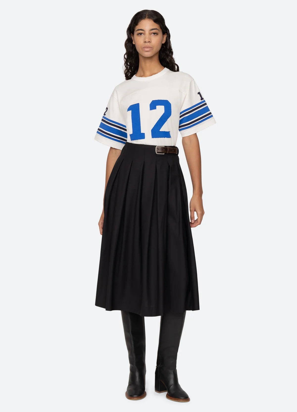 A woman in a relaxed-fit Finnley Football Graphic T-Shirt from Sea New York, featuring blue stripes and the number 12, paired with a black pleated skirt and tall black boots. She has long wavy hair and poses against a plain background, highlighting a sporty silhouette.