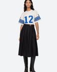 A woman in a relaxed-fit Finnley Football Graphic T-Shirt from Sea New York, featuring blue stripes and the number 12, paired with a black pleated skirt and tall black boots. She has long wavy hair and poses against a plain background, highlighting a sporty silhouette.