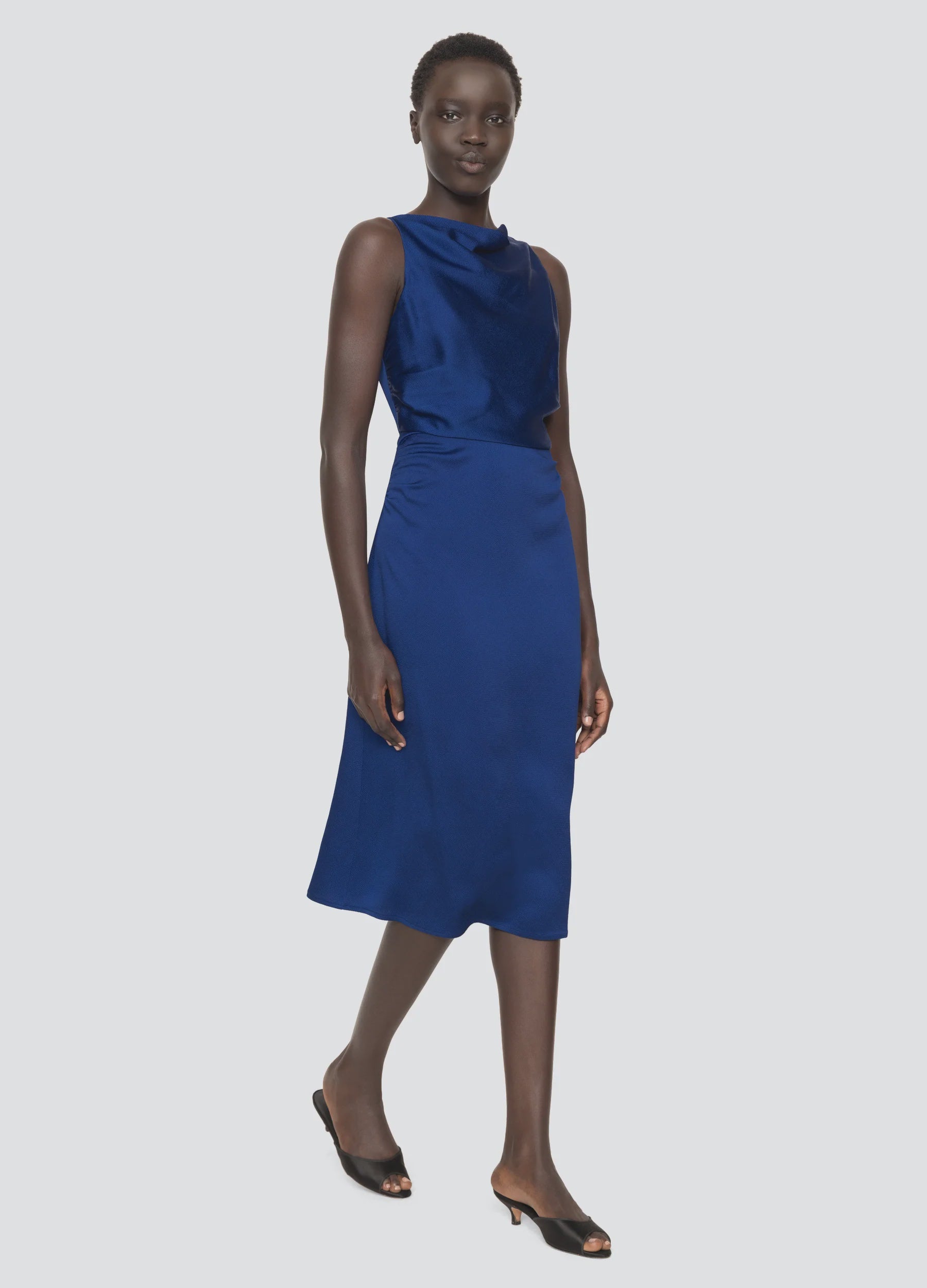 A person wearing the Noa Satin Halter Cowl Dress by Sea New York—characterized by its sleeveless design, knee-length cut, cobalt blue satin fabric, and cowl neckline—stands against a plain light background in black open-toe heels, looking forward.