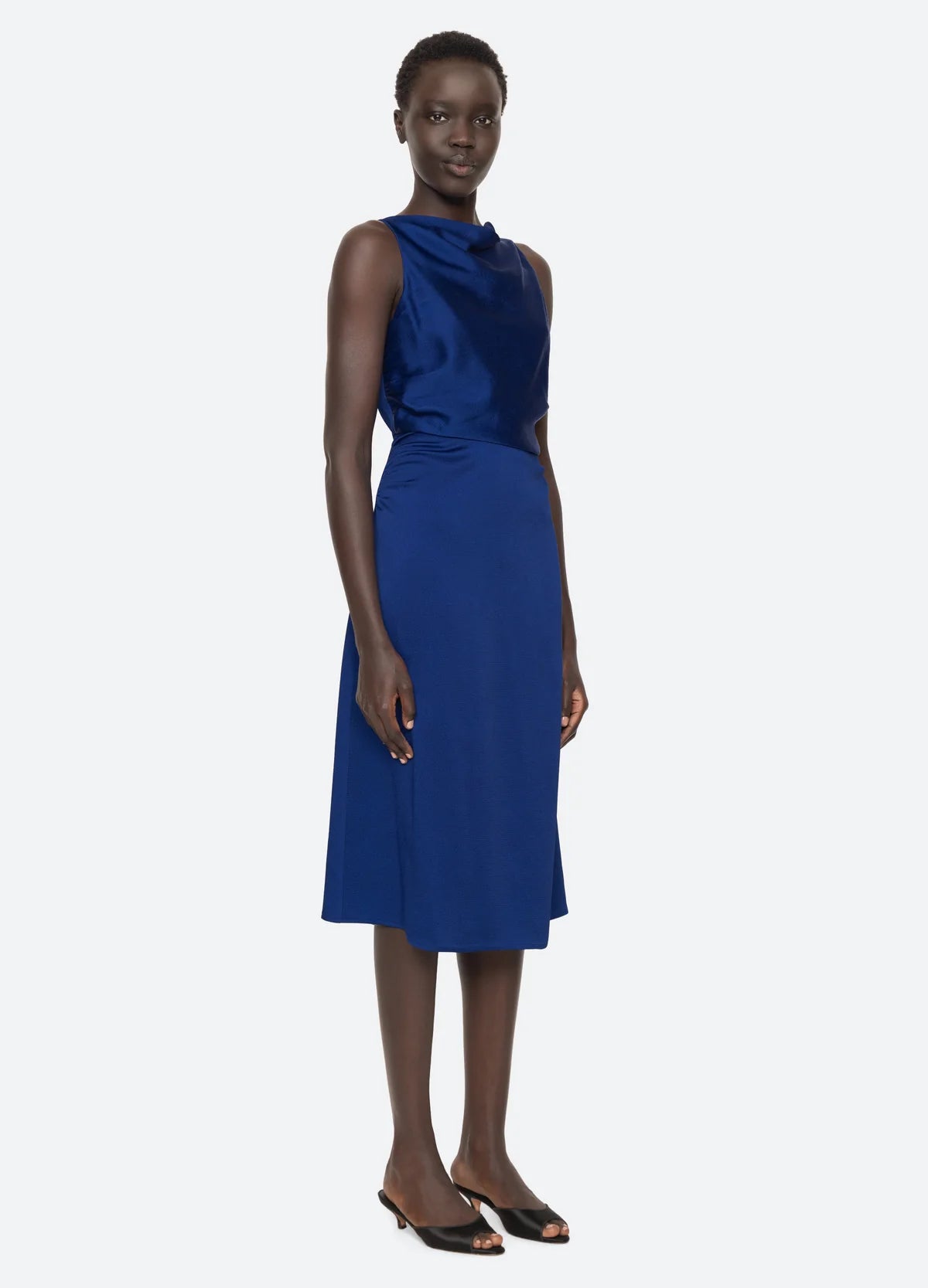 A person wearing the Noa Satin Halter Cowl Dress by Sea New York stands against a plain white background. The sleeveless, cobalt blue knee-length dress showcases ruched detailing, a high neckline, and a fitted waist. They pair it with black open-toed shoes and have short curly hair and a calm expression.