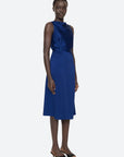 A person wearing the Noa Satin Halter Cowl Dress by Sea New York stands against a plain white background. The sleeveless, cobalt blue knee-length dress showcases ruched detailing, a high neckline, and a fitted waist. They pair it with black open-toed shoes and have short curly hair and a calm expression.