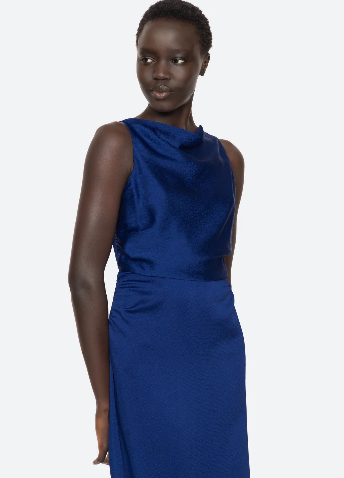 A person wearing the Noa Satin Halter Cowl Dress by Sea New York strikes a pose against a light gray backdrop. The sleeveless, cobalt blue dress showcases an elegant fitted waist and cowl neckline. Their short black hair is styled as they look slightly to the side.