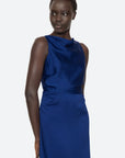 A person wearing the Noa Satin Halter Cowl Dress by Sea New York strikes a pose against a light gray backdrop. The sleeveless, cobalt blue dress showcases an elegant fitted waist and cowl neckline. Their short black hair is styled as they look slightly to the side.