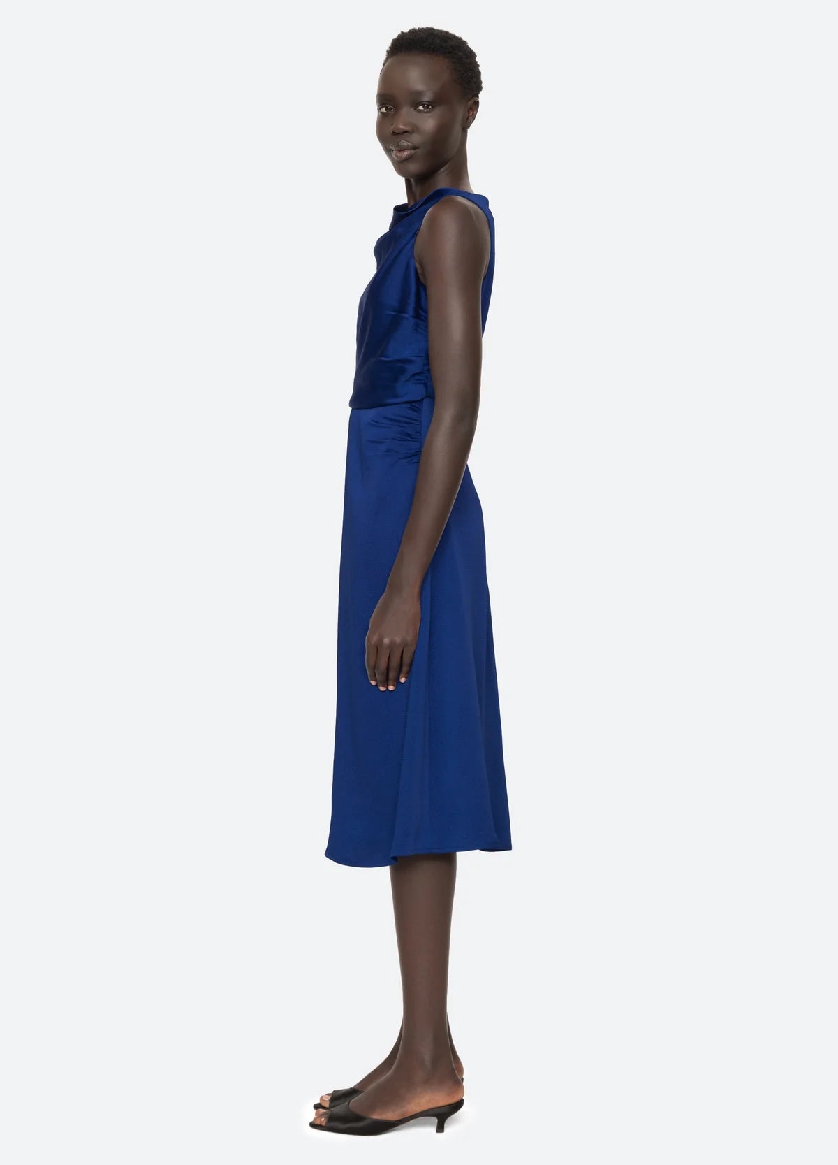 A person wearing the Sea New York Noa Satin Halter Cowl Dress, a sleeveless, knee-length dress in cobalt blue with ruched detailing, stands sideways while looking towards the camera. They are sporting black open-toe heels against a plain, light-colored background.