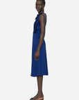 A person wearing the Sea New York Noa Satin Halter Cowl Dress, a sleeveless, knee-length dress in cobalt blue with ruched detailing, stands sideways while looking towards the camera. They are sporting black open-toe heels against a plain, light-colored background.