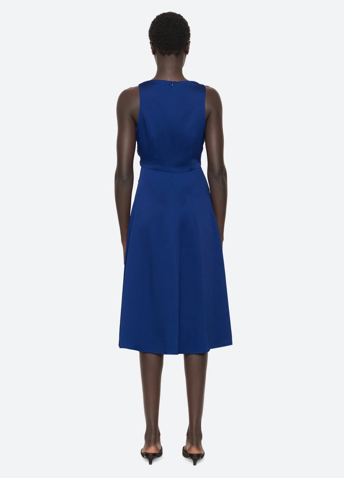 A person with short hair is wearing the Noa Satin Halter Cowl Dress, a sleeveless, knee-length cobalt blue garment from Sea New York, paired with black heels. The individual is standing facing away against a simple light background.