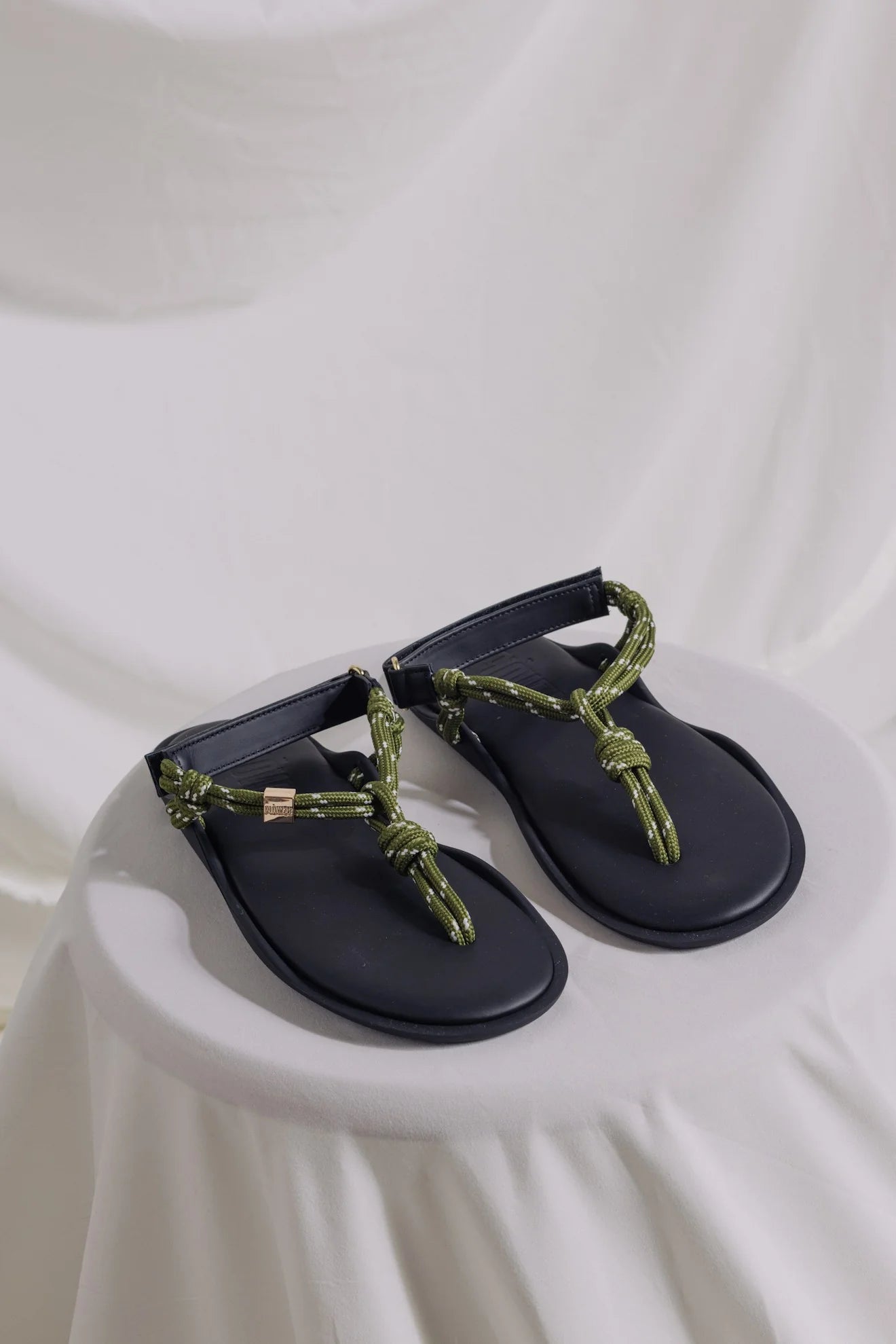 The FLO Sandals by Flowze, featuring black lightweight soles and green rope-style straps, are displayed on a round white fabric surface with a draped white cloth background.
