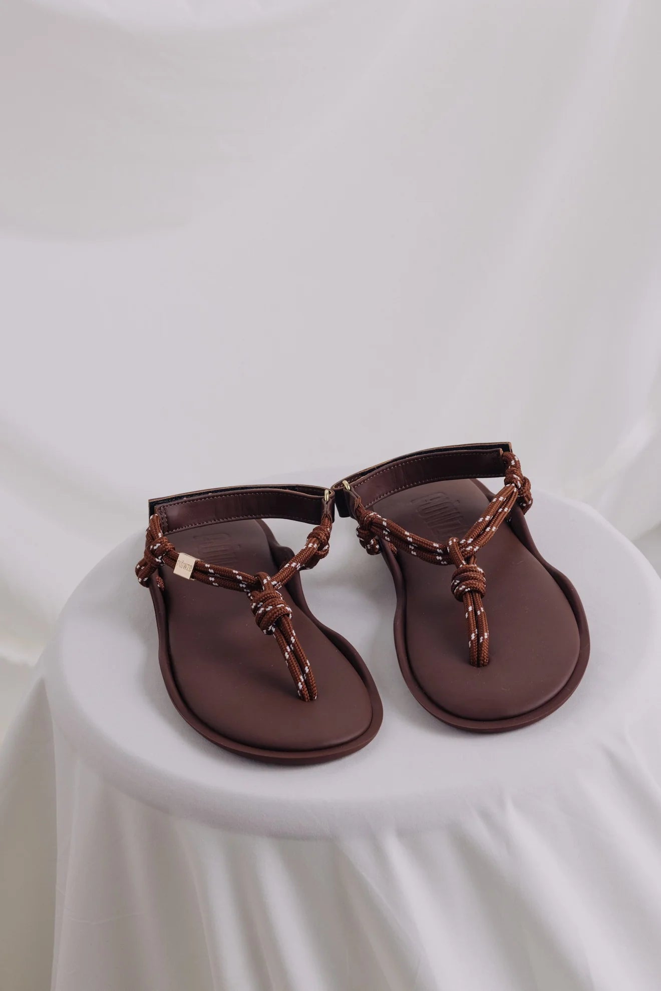 Flowze's FLO Sandals feature brown leather with knotted and braided straps, offering comfort. They are showcased on a round, white-draped table against a soft white backdrop.