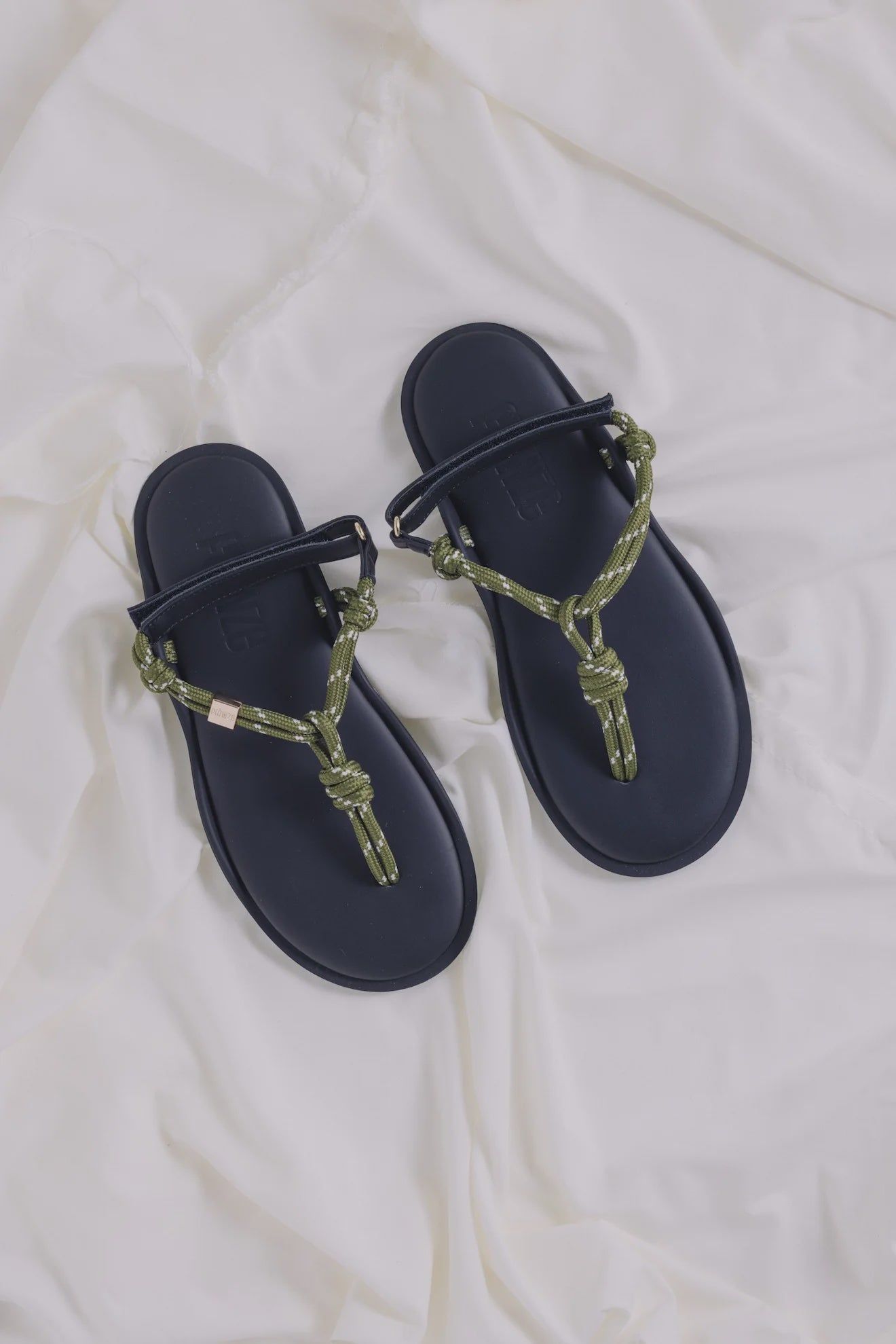 The Flowze FLO Sandals feature navy blue lightweight soles and green rope straps with adjustable knots, displayed on a soft white fabric background.