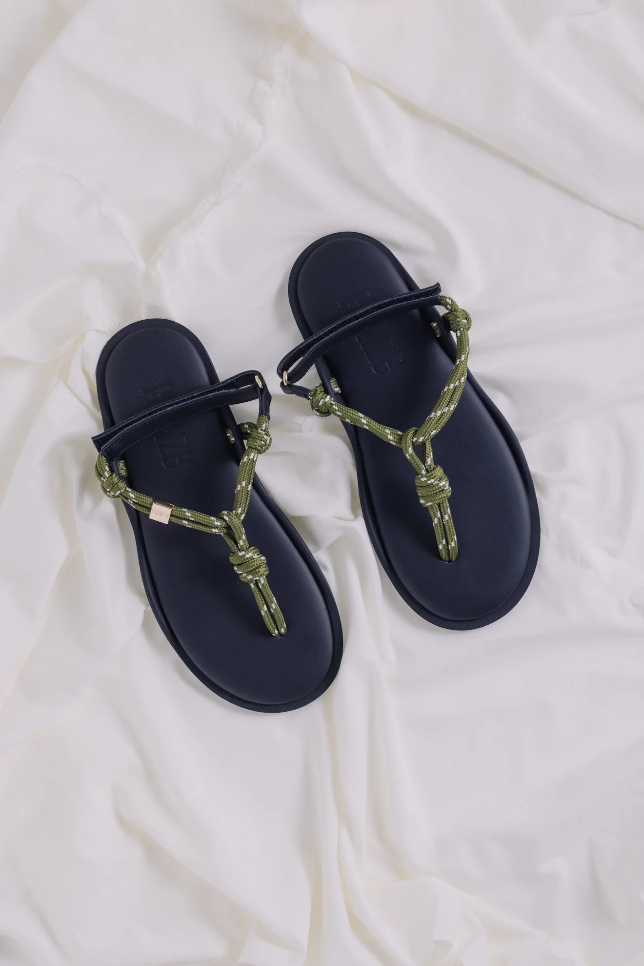 A pair of Flowze FLO Sandals, featuring black leather soles and green and beige knotted rope straps, rest on slightly wrinkled white fabric.
