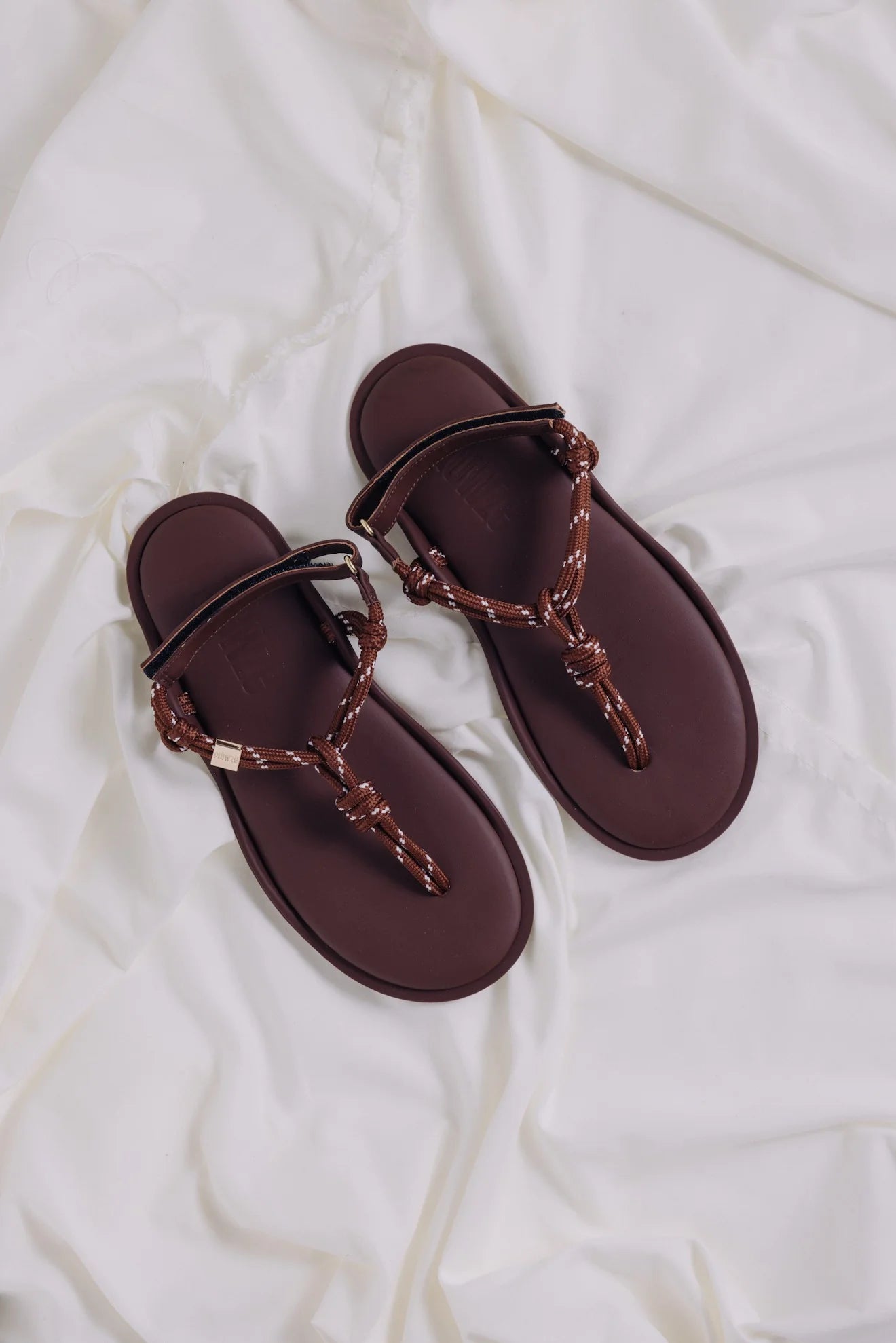 Flowze FLO Sandals—comfortable brown leather sandals with braided straps—are displayed on crumpled white fabric.