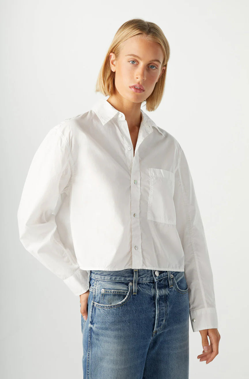 A person with short blonde hair wears the oversized white Ruth Crop Shirt by AMO Denim with blue jeans, standing against a plain light background with one hand in their pocket.