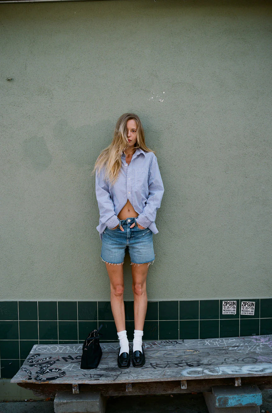 A person with long hair wearing the Ruth Oversized Shirt by AMO Denim and denim shorts stands against a green tiled wall. Their hands rest in their pockets, while a black bag sits on a distressed wooden bench in front of them.