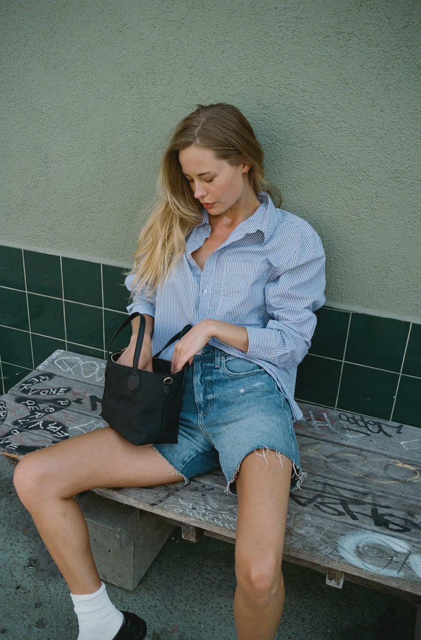 A woman with long blonde hair sits on a graffiti-covered bench, exuding vintage-inspired charm in her Ruth Oversized Shirt from AMO Denim and denim shorts. She holds a small black bag and looks downward, set against the backdrop of a green tiled wall.