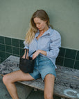 A woman with long blonde hair sits on a graffiti-covered bench, exuding vintage-inspired charm in her Ruth Oversized Shirt from AMO Denim and denim shorts. She holds a small black bag and looks downward, set against the backdrop of a green tiled wall.