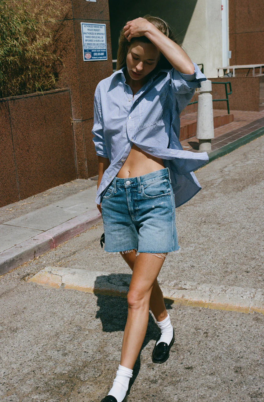 Wearing the Ruth Oversized Shirt by AMO Denim, an individual in a classic blue-striped design pairs it with vintage wash denim shorts as they walk down a sunny street, adjusting their hair. Their ensemble is completed with white socks and black shoes, set against the backdrop of a brown wall and sign.