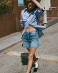 Wearing the Ruth Oversized Shirt by AMO Denim, an individual in a classic blue-striped design pairs it with vintage wash denim shorts as they walk down a sunny street, adjusting their hair. Their ensemble is completed with white socks and black shoes, set against the backdrop of a brown wall and sign.