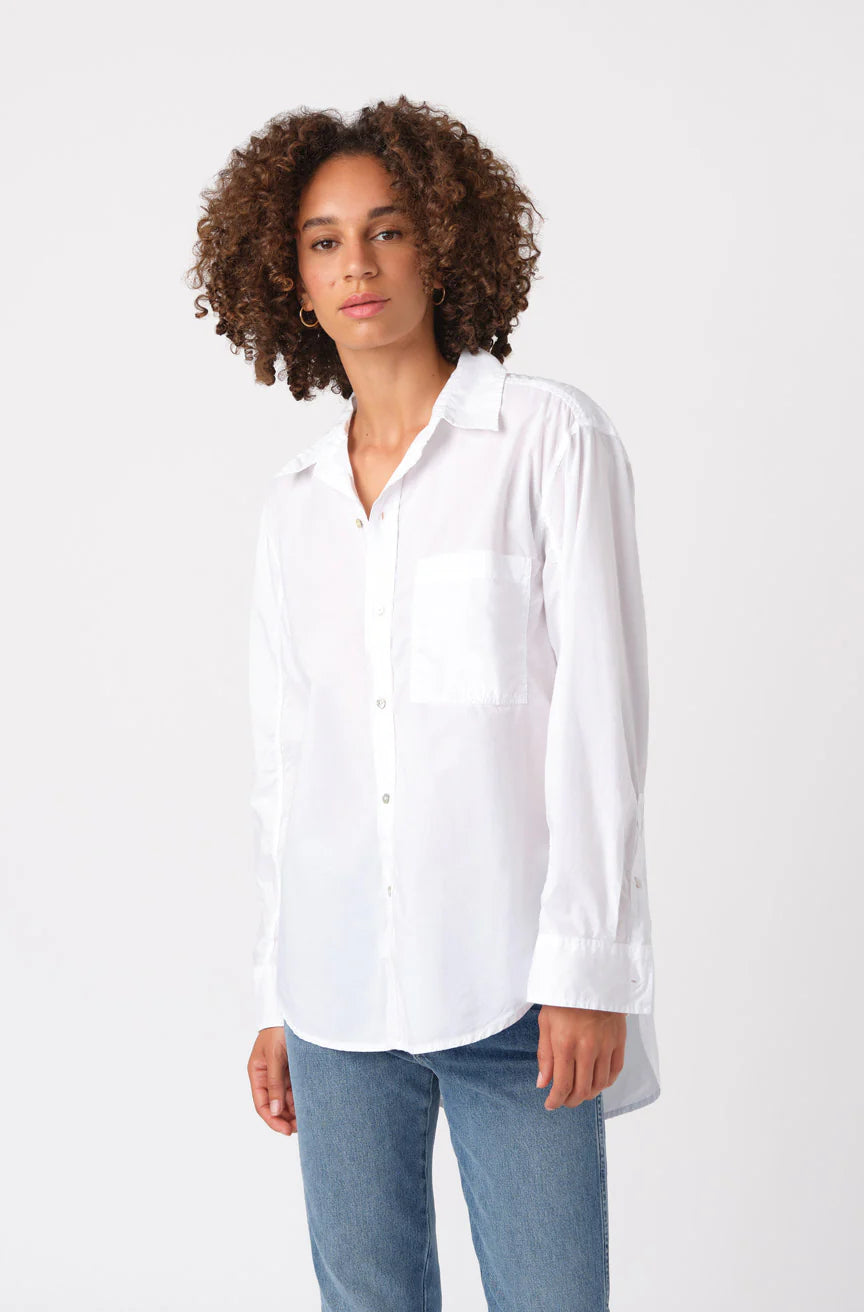 A person with curly hair models the AMO Denim Ruth Oversized Shirt and blue jeans, standing against a plain white background while looking at the camera with a neutral expression.