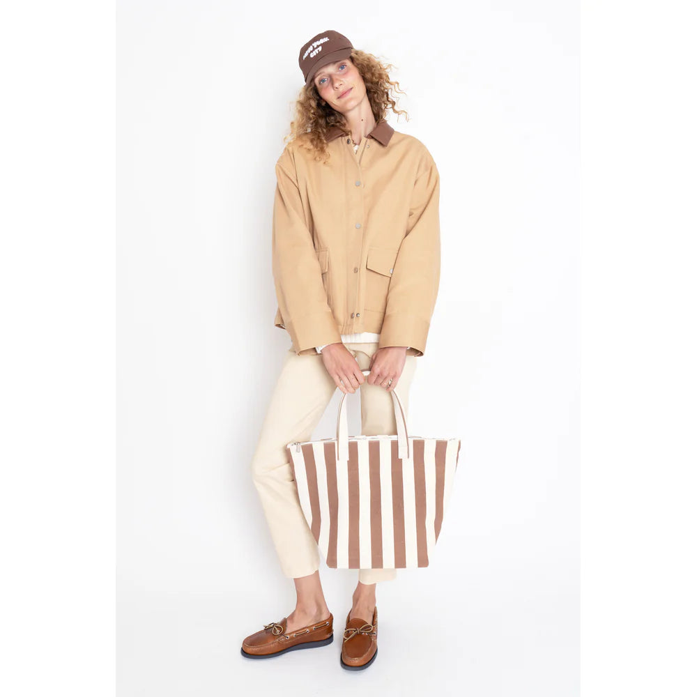 A person with curly hair and glasses stands against a plain white background, wearing a beige chore coat featuring a vegan leather collar, cream pants, brown shoes, and a brown cap. They're holding "The Ripley," a large brown and white striped tote by Kule.