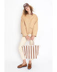 A person with curly hair and glasses stands against a plain white background, wearing a beige chore coat featuring a vegan leather collar, cream pants, brown shoes, and a brown cap. They're holding "The Ripley," a large brown and white striped tote by Kule.