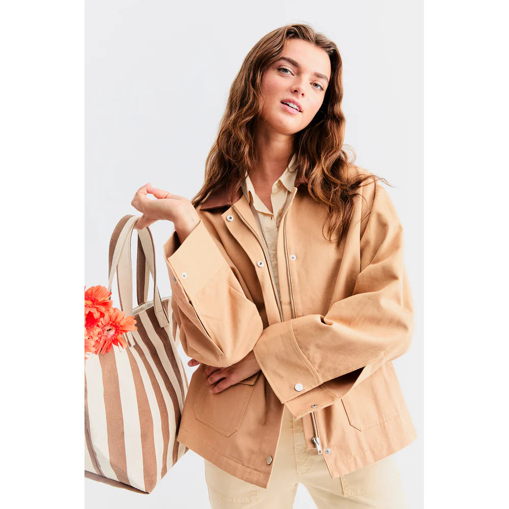 A person with long hair wears The Ripley, a tan chore coat by Kule made from a cotton-linen blend, over a beige shirt. They hold a striped tote bag filled with orange flowers against a plain white background.