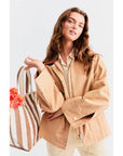 A person with long hair wears The Ripley, a tan chore coat by Kule made from a cotton-linen blend, over a beige shirt. They hold a striped tote bag filled with orange flowers against a plain white background.