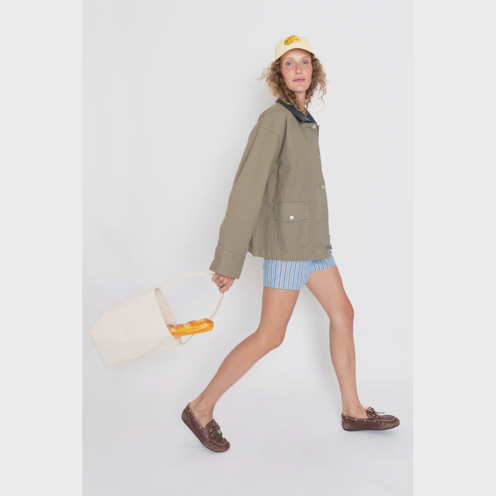 A person wears "The Ripley" by Kule—a beige chore coat with a vegan leather collar—paired with blue striped shorts, brown shoes, and a white cap featuring yellow text, while holding a baguette-filled white bag. They pose with one leg extended, gazing directly at the camera.