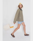 A person wears "The Ripley" by Kule—a beige chore coat with a vegan leather collar—paired with blue striped shorts, brown shoes, and a white cap featuring yellow text, while holding a baguette-filled white bag. They pose with one leg extended, gazing directly at the camera.