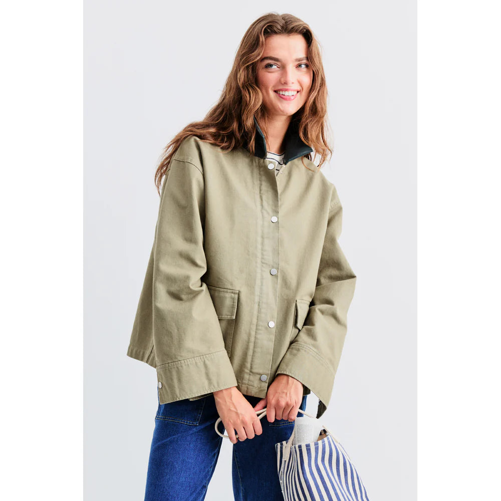 A person with long hair is wearing The Ripley, a khaki chore coat by Kule with front pockets, a vegan leather collar, and silver buttons. They're smiling while holding a striped tote against a plain light gray background, paired with blue jeans.