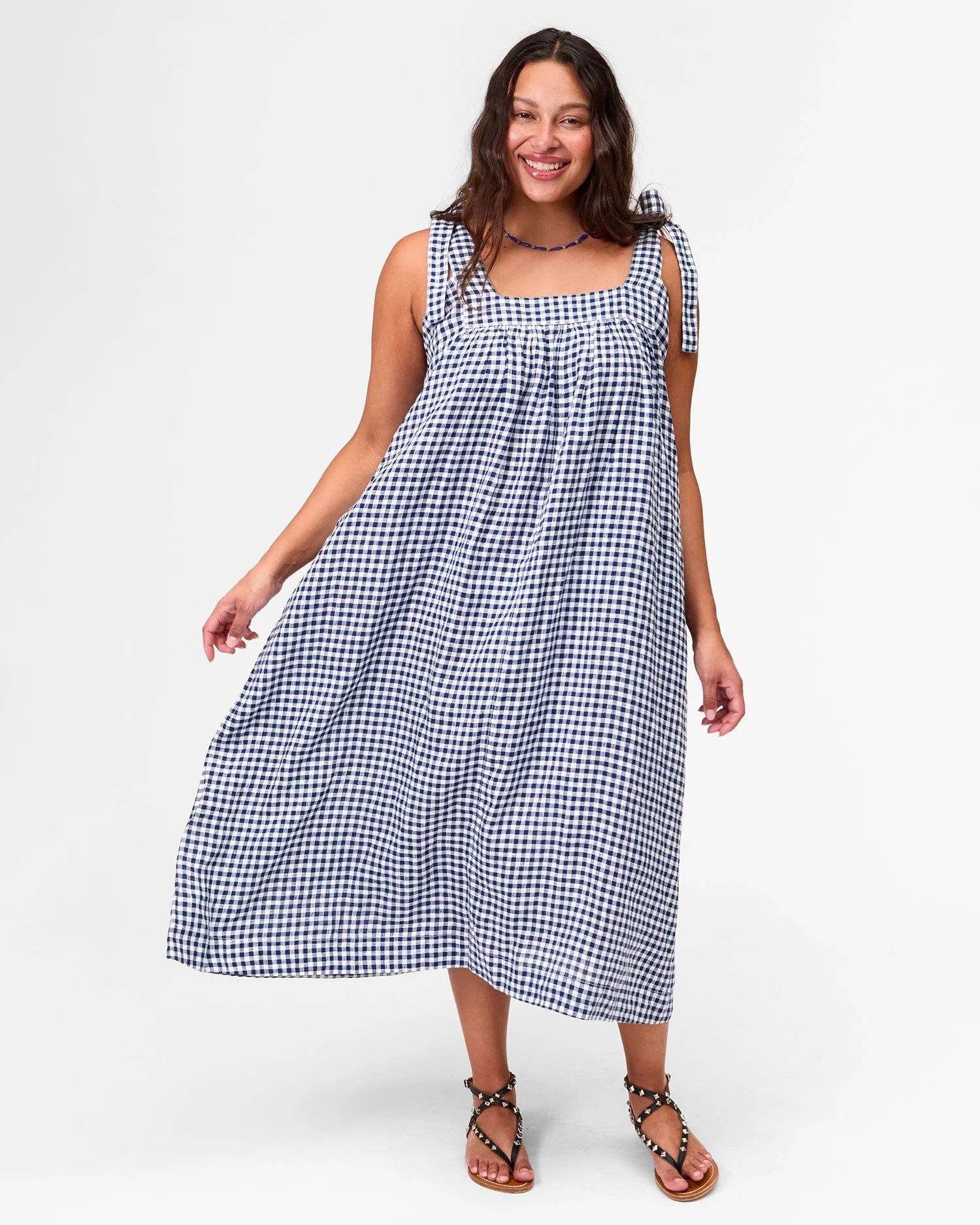 A woman smiles in the Rosalie Dress by Clare Vivier, a loose, sleeveless blue-and-white checkered linen dress with shoulder ties and pockets. She pairs it with sandals and poses against a plain white background.