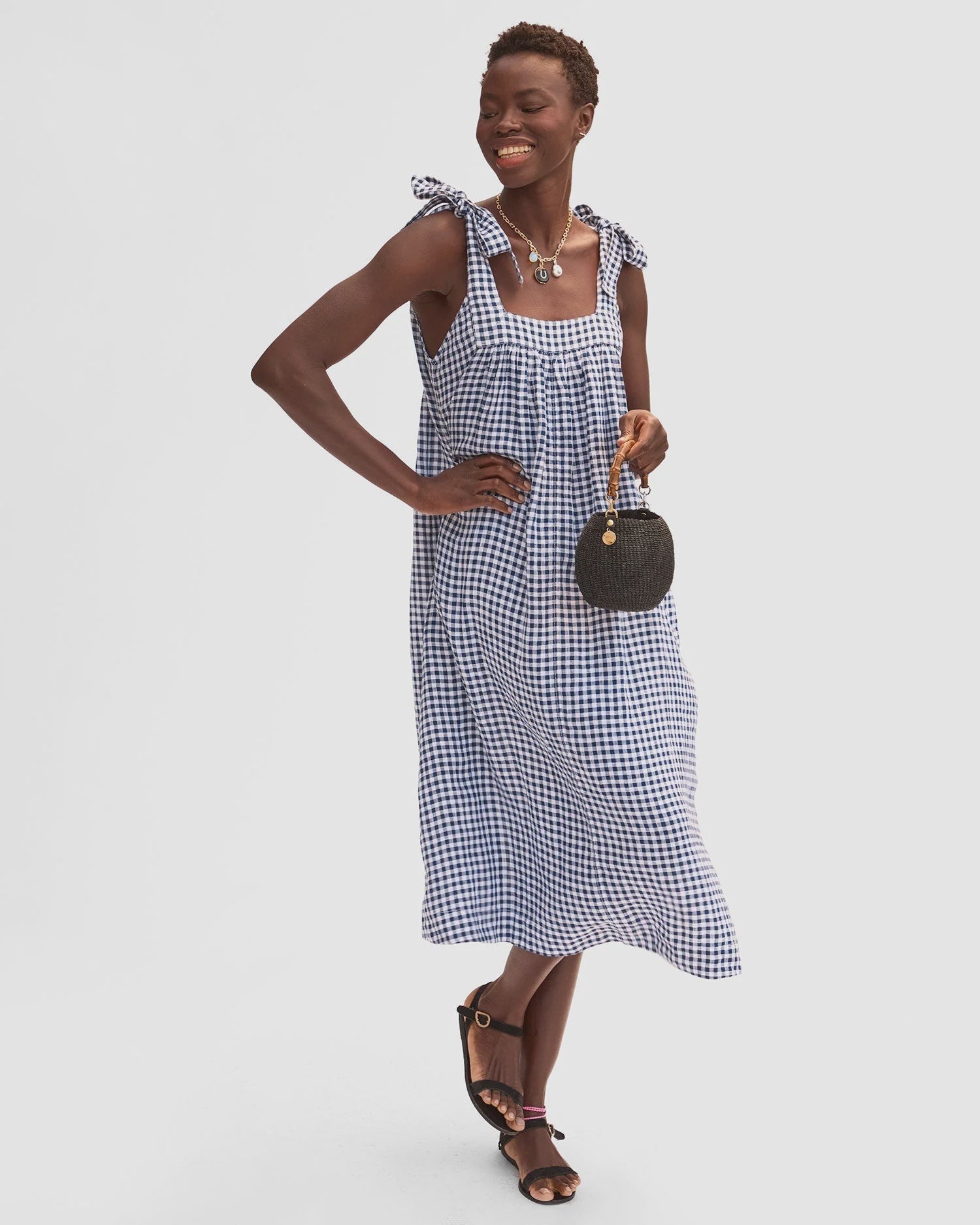 A smiling woman wears the Rosalie Dress by Clare Vivier, a blue and white checkered linen dress with a square neckline and tie straps, styled with black sandals and a small round black handbag, posing against a plain backdrop.