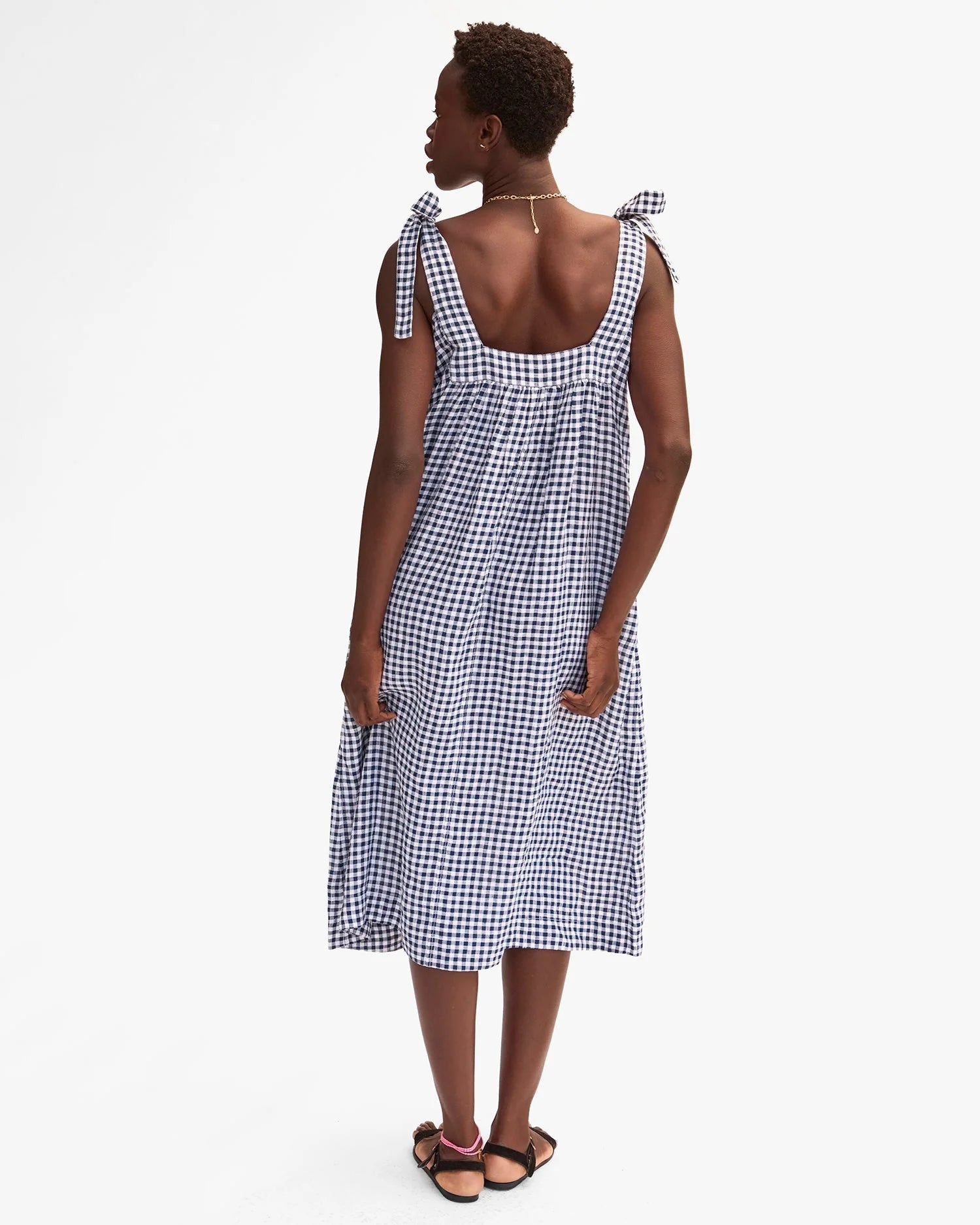 A woman with short hair and a gold necklace wears the Clare Vivier Rosalie Dress, a sleeveless linen dress with black and white gingham pattern and tied shoulder straps, paired with black sandals. She stands against a plain white background.